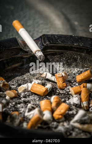 Eine brennende Zigarette mit langen Asche befindet sich auf der Seite eine vollständige und sehr dreckig Aschenbecher voller Zigarettenkippen. Stockfoto