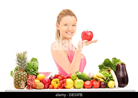 Junge, lächelnde weibliche hält einen roten Apfel und posiert hinter einem Tisch voller Gemüse und Obst isoliert gegen weiße staatlich Stockfoto