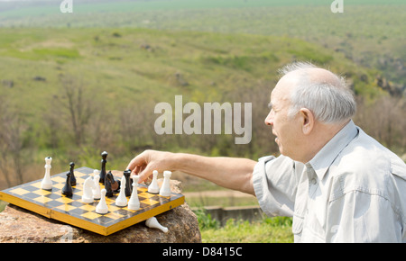 Pensionierter Mann spielt Schach auf dem Land erreichen über eine Spielmarke greifen zu bewegen gegen einen schönen Talblick Stockfoto