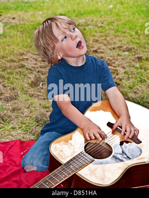 Junge ist ein aufstrebender Musiker, wie er singt ein Lied, während Zupfen einer Gitarre, die in der Nähe von ihm zu legen ist. Er befindet sich im freien Stockfoto