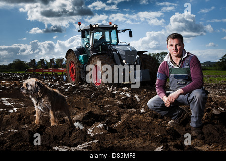 Porträt eines jungen Landwirts und seinem kleinen Hund vor einem Traktor und Pflug in einem Acker. Stockfoto