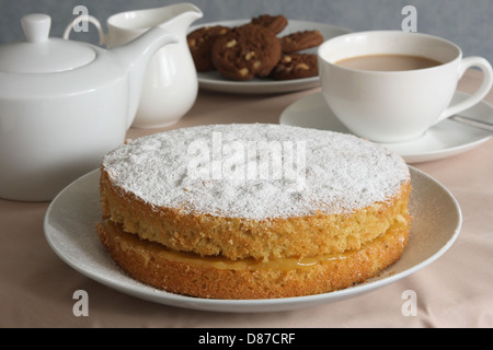 Nachmittags-Tee. Eine frisch gebackene Biskuit dominiert das Foto, eine Teekanne, Kanne, eine Tasse Tee und Kekse sind im Hintergrund. Stockfoto