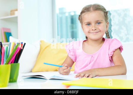 Niedliche kleine Mädchen vom Tisch mit Buntstiften und offene Copybook vor ihr sitzt Stockfoto