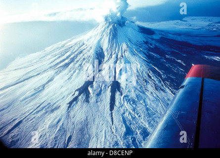 Der Augustine-Vulkan in Alaska ist ein aktiver Stratovulkan, der für seine explosiven Eruptionen bekannt ist. Dieses Foto zeigt den imposanten Berggipfel mit Rauchwolken, die vom Gipfel aufsteigen und auf seine vulkanische Aktivität hinweisen. Die umliegende Landschaft umfasst schneebedeckte Berge und zerklüftetes Gelände, das typisch für die Region ist. Stockfoto