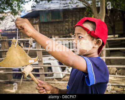 25. Mai 2013 - Mae Sot, Tak, Thailand - Ringe ein burmesischer jungen einen Gebet Gong kündigt die Ankunft des burmesischen Mönch und Novizen in einem inoffiziellen Dorf der birmanischen Flüchtlinge nördlich von Mae Sot, Thailand. Ihre Gemeinde ist etwa 50 Meter von der burmesischen Grenze zu Thailand. Etwa 200 Menschen leben in strohgedeckten Hütten in der gesamten Gemeinschaft zu verbreiten. Sie sind nah genug, um Mae Sot, die einige können in der Stadt arbeiten und birmanische Händler von Mae Sot kommen, um ihr Dorf, um mit ihnen Geschäfte machen. Die Birmanen im Dorf nicht Residency Papiere für Thailand und gelten als technisch illegal immigr Stockfoto