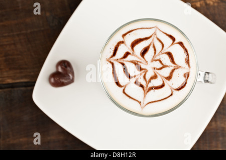 Ein Blick von oben auf eine Glaskaffeetasse gefüllt mit einem Latte mit komplizierter Latte-Kunst im rosetta-Stil, hergestellt mit Schokoladensirup auf cremigem Milchschaum. Der Becher sitzt auf einer weißen quadratischen Platte Stockfoto
