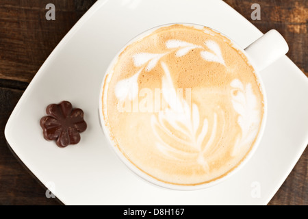 Blick von oben auf eine weiße Keramiktasse mit Kaffee und komplizierten Latte Art, einschließlich Blatt- und Blumenmustern im Milchschaum. Der Becher liegt auf einer quadratischen weißen Untertasse, die auf einer dunklen Untertasse steht Stockfoto