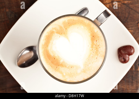Ein Blick von oben auf einen Latte in einem klaren Glasbecher mit Metallgriff und einem herzförmigen Design im Milchschaum. Der Becher sitzt auf einer weißen quadratischen Untertasse, begleitet von einem Metalllöffel links Stockfoto