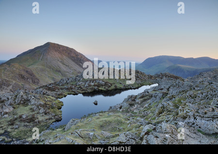 Kleinen Teich auf dem Gipfel des Heuhaufen in der Morgendämmerung im englischen Lake District, mit hohen Felsen im Hintergrund Stockfoto