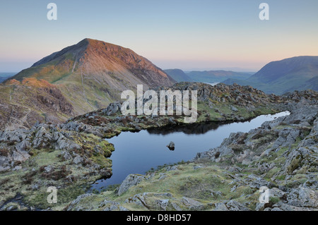 Kleinen Teich auf dem Gipfel des Heuhaufen in der Morgendämmerung im englischen Lake District, mit hohen Felsen im Hintergrund Stockfoto