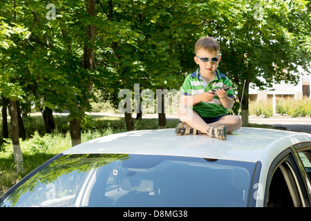 Blonde junge moderne modische Sonnenbrille sitzt auf der Oberseite ein weißes Auto spielen, mit grün belaubten Bäume in der Stockfoto