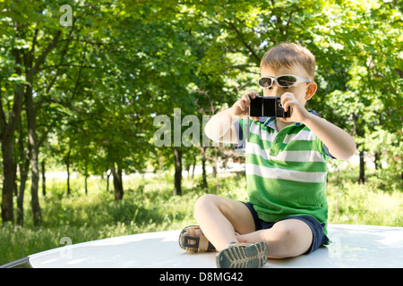 Blonde Junge sitzt auf der Oberseite ein weißes Auto mit gekreuzten Beinen, Fotografieren mit dem Handy Stockfoto