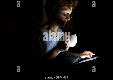 Frau, die nicht schlafen, sitzen im Bett, Tee trinken und mit Hilfe eines Tablet-Computers Stockfoto