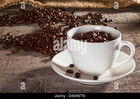 Kaffeebohnen und rustikalen Hintergrund - Kaffee-Tasse und Untertasse gefüllt mit Kaffeebohnen, Kaffeebohnen mit Leinensack hinter... Stockfoto