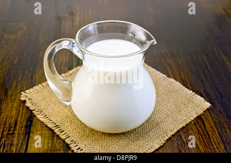 Milch in eine Glaskanne auf einer Serviette aus Sackleinen auf dunklem Hintergrund Holzbrett Stockfoto