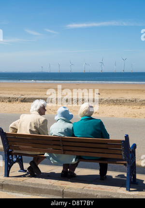 Drei ältere Frauen sitzen auf Bank in der Nähe von Strand in Redcar, Cleveland, England, UK. Teesside Offshore-Windpark im Hintergrund Stockfoto