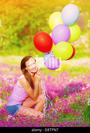 Hübsches Mädchen sitzen auf rosa Blumen Wiese und hält in Händen bunte Luftballons, Spaß im Freien, Sommerurlaub Stockfoto