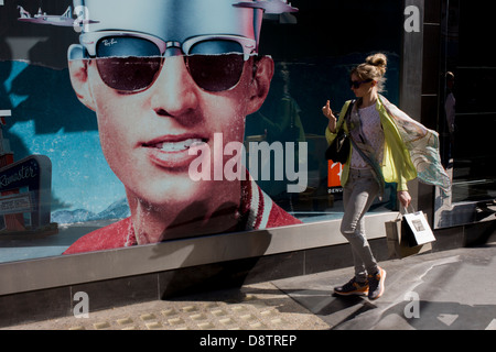 Eine trendige junge Frau geht-durch ein Plakat eines Modells zeigt stilvolle Farben außerhalb einer Sonnenbrille Schaufenster verkauft Ray Bans auf Long Acre im Londoner Covent Garden. Stockfoto