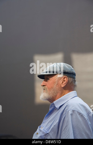 Mann der 50er Jahre, mit grauen Bart tragen Tuch Mütze und blaues Hemd, graue Wand mit Flecken von Sonnenlicht, im Profil Stockfoto