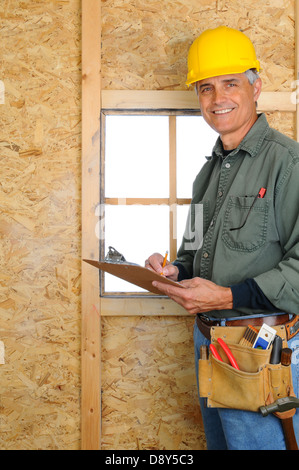 Einen mittleren Alters Auftragnehmer stehen im Neubau auf einem Clip Board schreiben. Mann trägt Jeans, Arbeitshemd, Werkzeuggürtel. Stockfoto