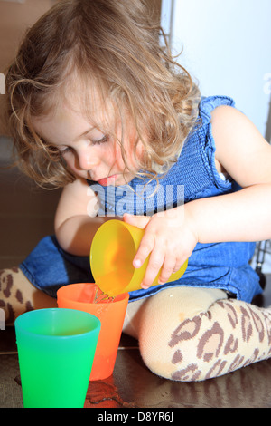 2 Jahre altes Mädchen lernen und spielen Wasser aus einem farbigen Becher in einen anderen Stockfoto