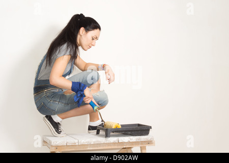 Frau mit einer Walze, um eine Wand zu malen, während der Sanierung oder Renovierung ihres Hauses malen aus einem Plastikbehälter nehmen Stockfoto