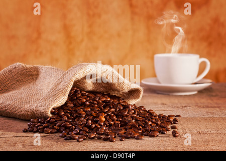 Kaffeebohnen und Leinensack - gerösteten Kaffeebohnen, die aus einem Leinensack, Tasse Kaffee mit sichtbaren Dampf verschütten... Stockfoto