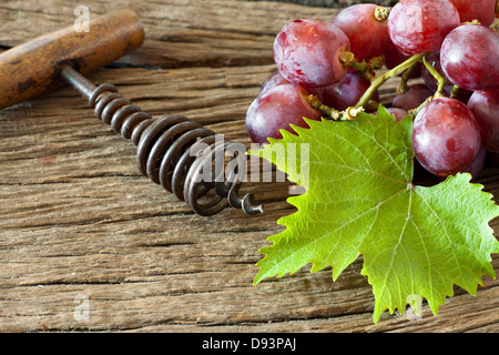 Alter Korkenzieher mit Holzgriff liegt zwischen roten Trauben und Weinblättern auf rustikalen hölzernen Hintergrund Stockfoto