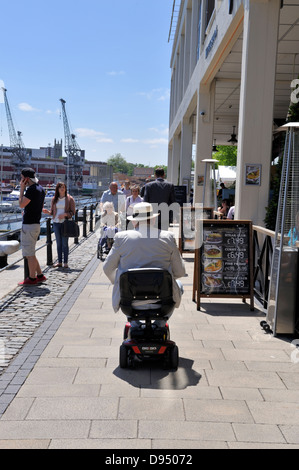 Älterer Mann fahren e-Mobilität Scooter entlang Bristol Harbourside Waterfront Gehweg, UK Stockfoto