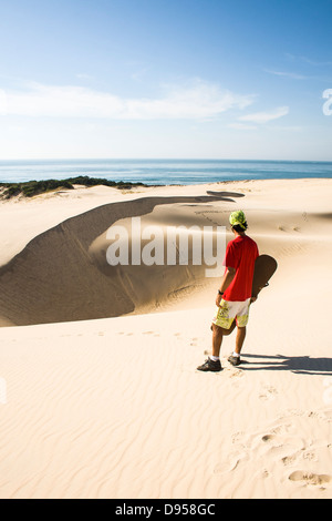 Rückansicht eines erwachsenen Mannes hält ein Sandboard und Blick auf das Meer auf den Dünen von Siriu Beach. Stockfoto