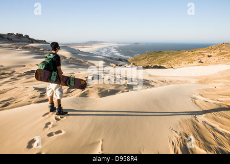 Rückansicht eines jungen Mannes hält ein Sandboard und Blick auf das Meer auf den Dünen von Cape Santa Marta. Stockfoto