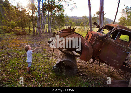 Kleine Junge steht durch die rostenden Kabine von einem alten LKW gestützt auf einem Baumstamm in den australischen Busch Stockfoto