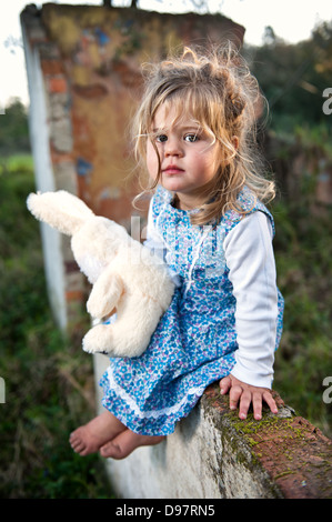 Junge Kleinkind mit langen blonden Haaren und blumigen Kleid sitzt auf einer Mauer der alten baufälligen Gebäude, einen Teddybär. Stockfoto