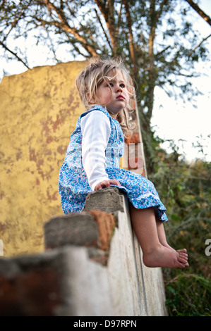 Blonde Kleinkind mit blauen Blumen Kleid und barfuß, auf eine alte verfallene Mauer starrte emotionslos sitzen. Stockfoto