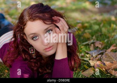 Schöne junge Frau mit lockigem Haar liegt am Bauch Blatt bedeckt Gras, einerseits an Wange, Blick in die Kamera Stockfoto