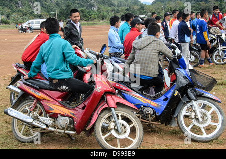 Studenten-Zuschauer Scooters Crossbow Contest Vieng XAI Laos // VIENG XAI, Laos — Studenten-Zuschauer stellen sich auf ihren Scootern und Motorrädern an, um einen Armbrustwettbewerb auf einem unbefestigten Spielfeld in der Nähe von Vieng XAI im Nordosten von Laos zu beobachten. Die traditionelle Sportveranstaltung zieht Teilnehmer und Zuschauer aus den umliegenden Dörfern dieser ländlichen Bergregion an. Vieng XAI, in der Provinz Houaphanh gelegen, ist bekannt für seine Kalksteinkarstlandschaft und das Höhlennetz, das während des Zweiten Indochina-Krieges als Hauptquartier für die Pathet Lao diente. Die Stadt liegt in der Nähe des Bor Stockfoto