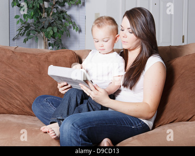 Hübsche junge kaukasischen Mutter und Sohn blondes Kleinkind sitzt zu Hause auf Sofa zusammen auf einem drahtlosen mobilen Tablet lesen. Stockfoto