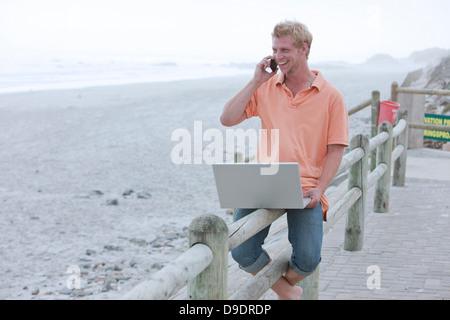 Mann sitzt am Strand Geländer mit Handy und laptop Stockfoto