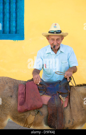 Porträt der Greis rauchende Zigarre, posiert vor einer gelben Wand mit seinem Esel für touristische Pesos, Trinidad, Kuba, West Indies Stockfoto