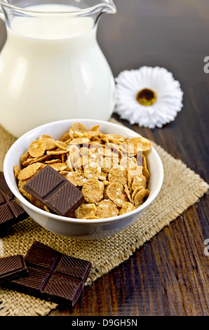 Corn Flakes mit Scheiben von Schokolade, Milch im Glaskrug, hessischen Tuch, weiße Blume auf einem Holzbrett Stockfoto