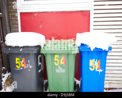 Wheelie-Kästen vor einem Haus, mit Schnee bedeckt. Schwarz, grün und blau Kästen. Stockfoto