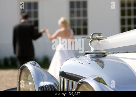 Detail der klassischen Hochzeitsauto mit Emblem und Scheinwerfer mit Braut und Bräutigam im Hintergrund Hand in Hand Stockfoto
