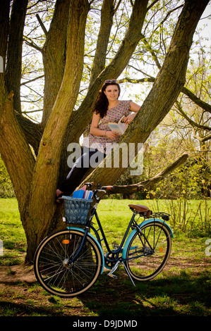 Junge kaukasische Frau, die auf einem Baum sitzt und sich mit einem Buch entspannt. Fahrrad unten geparkt. Stockfoto