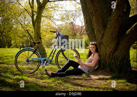Junge kaukasische Frau sitzt unter einem Baum und entspannt ein Buch zu lesen. Stockfoto