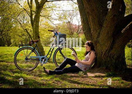 Junge kaukasische Frau sitzt unter einem Baum und entspannt ein Buch zu lesen. Stockfoto