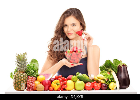 Junge, lächelnde weibliche hält eine Schale mit Erdbeeren und posiert hinter einem Tisch voller Gemüse und Obst Stockfoto