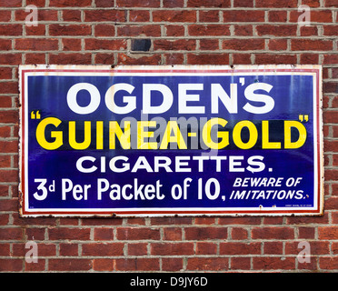 Vintage blau, weiß und gelb Emailleschild Werbung Ogdens Guinea Gold Zigaretten, auf Ziegelmauer Stockfoto