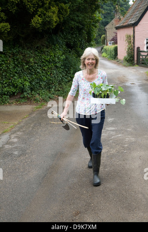 Frau trägt Sämlinge und Gartenarbeit Werkzeuge in einer Dorfstraße Stockfoto