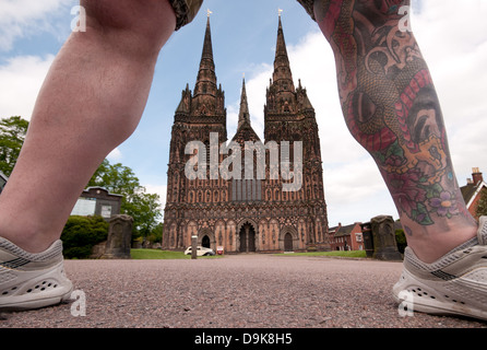 Ungewohnter Anblick der Westfassade der Kathedrale von Lichfield gesehen durch die tätowierten gut bemuskelte Beine des Mannes in kurzen Hosen Stockfoto