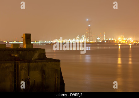 Öl-Raffinerie mit hohen Flare Stacks in der Nacht mit einem schmutzigen orangefarbenen Himmel und Licht, ein Dock im Vordergrund. Stockfoto